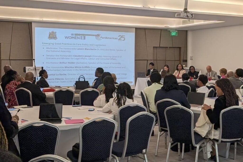 Panel discussion at a conference room with speakers seated at a long table near a projection screen displaying 'WOMEN' and event details; attendees sit at round tables with laptops and notebooks nearby.