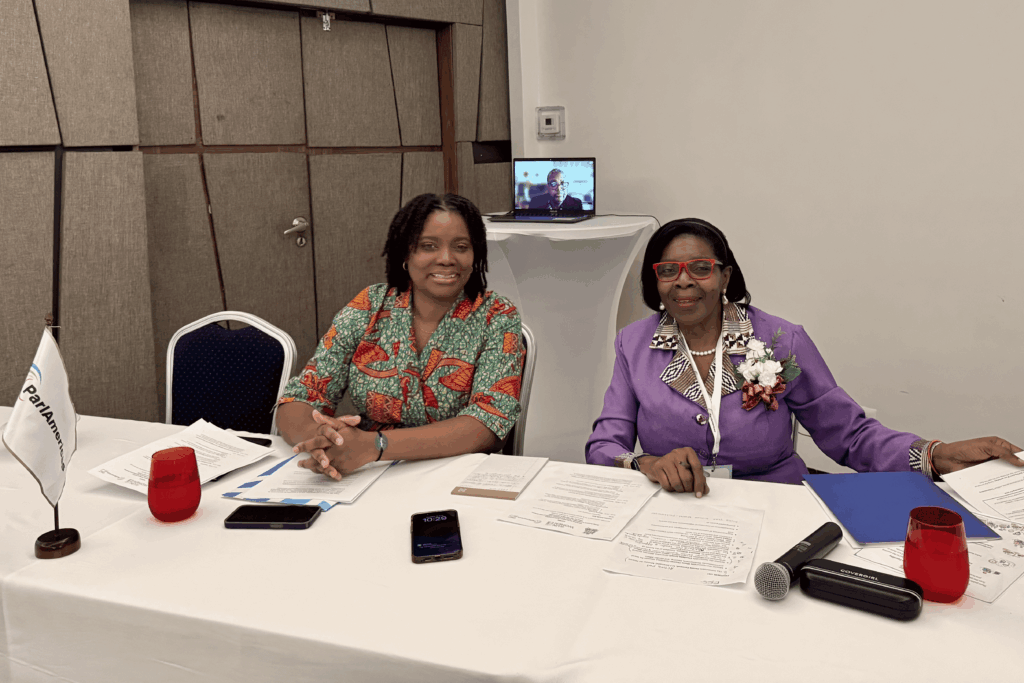 Two women sit at a table covered with documents; a laptop on a stand behind them shows a person in a video call as they pose for the photo.