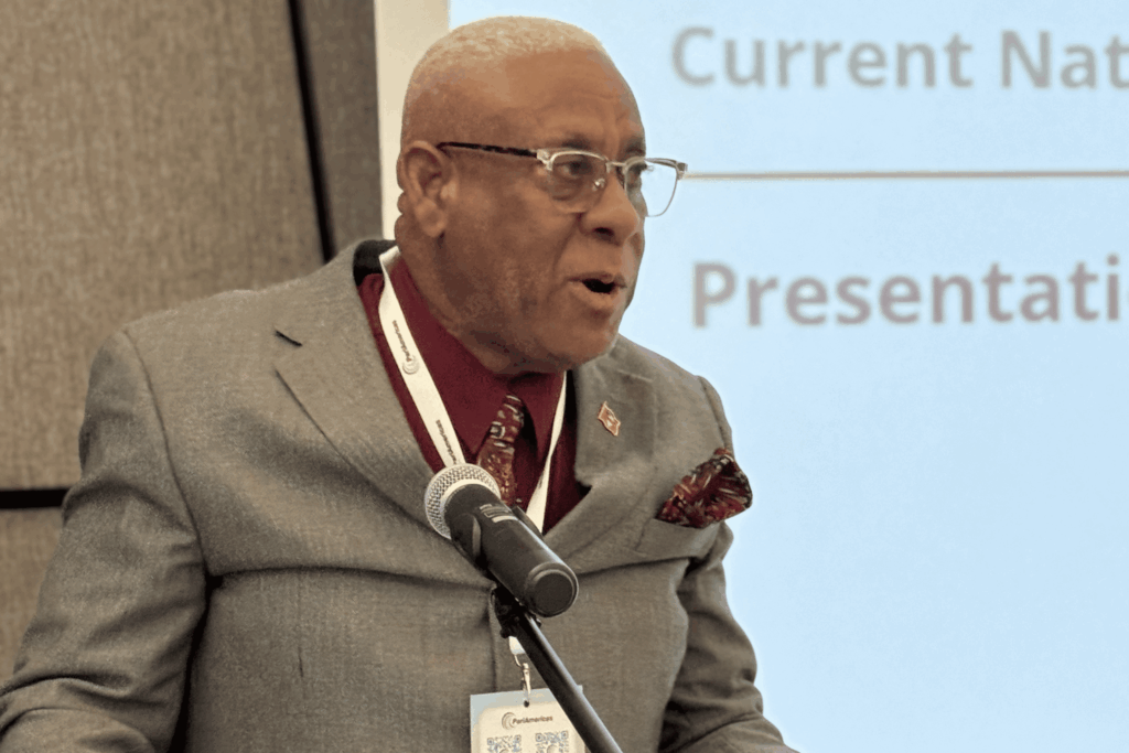 Speaker in a gray suit at a podium, wearing a lanyard and microphone in front of a light blue backdrop.