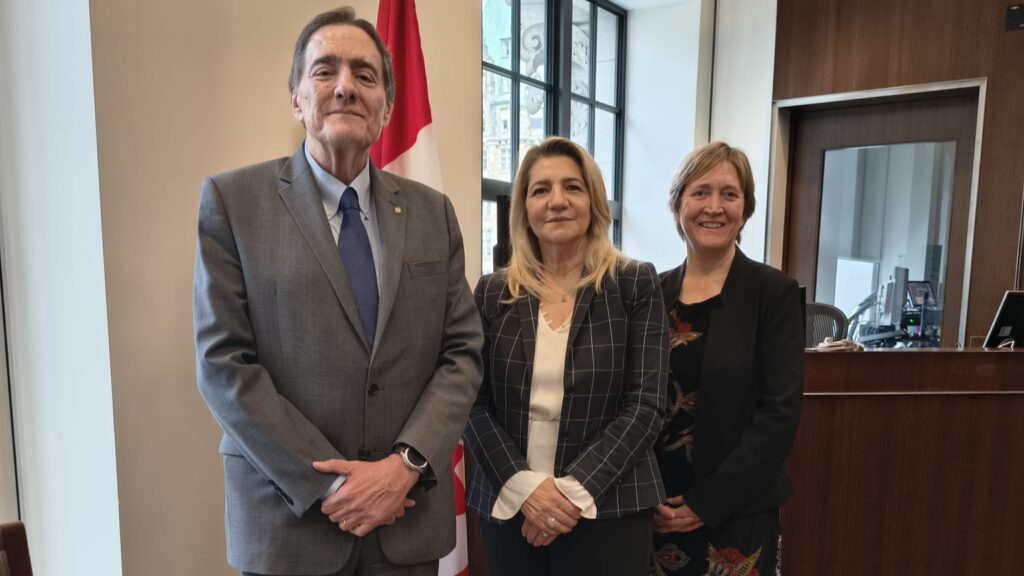 Three professionals pose for a photo in a office lobby with a flag in the background, near a reception desk to the right.