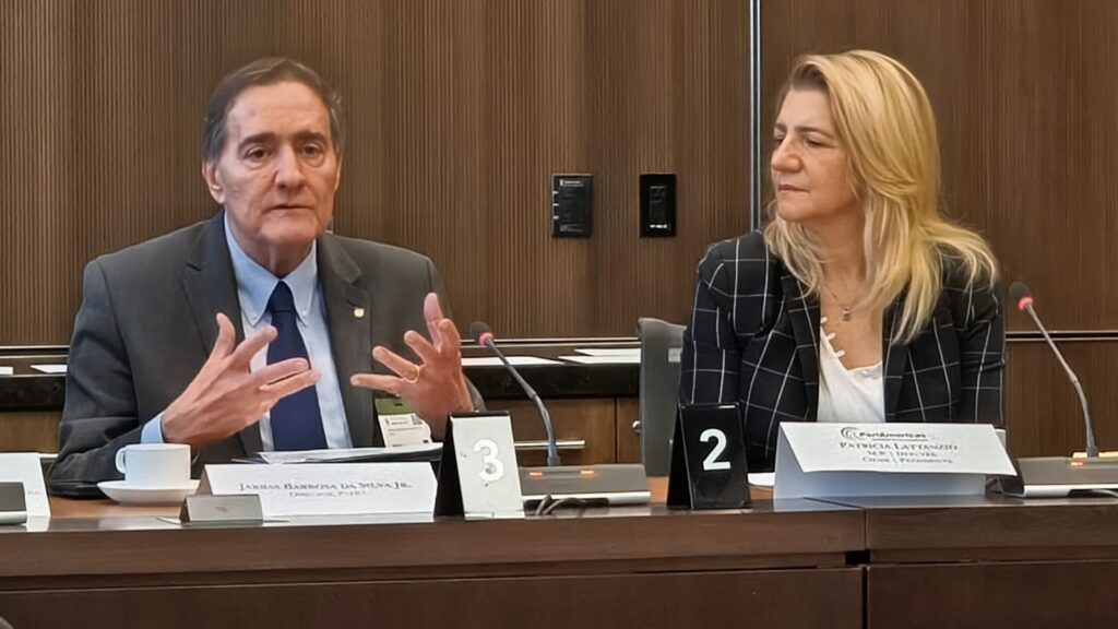 Two panelists seated at a wood-paneled table, man speaking with hand gestures while a woman listens nearby, microphones in use.