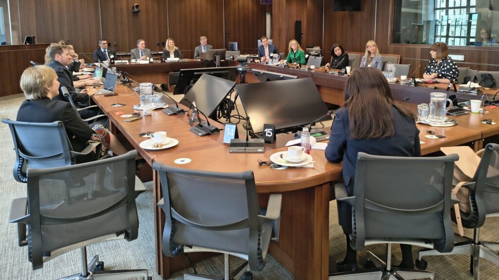 Professionals seated around a large U-shaped conference table in a formal meeting room, with laptops and documents visible.