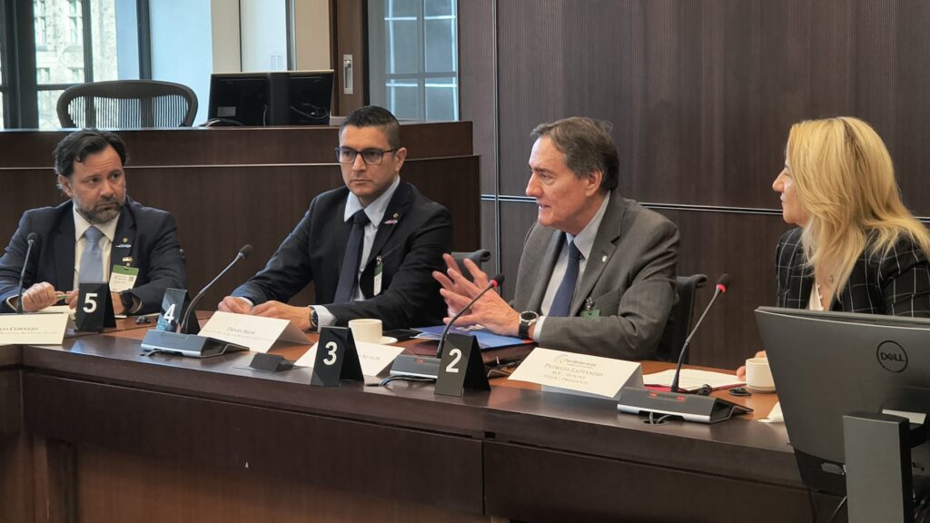 Four panelists in suits seated at a long conference table, speaking into microphones during a formal panel discussion, with numbered name placards in front (2–5).