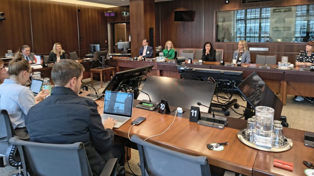 Panel of people seated around a large wood-paneled conference table with laptops, microphones, and nameplates in a formal meeting room.