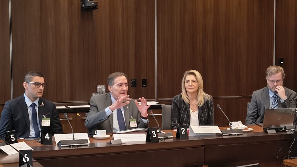 Panel of five people seated at a long conference table, with name placards 4–5 visible, discussing a topic in a formal meeting room.