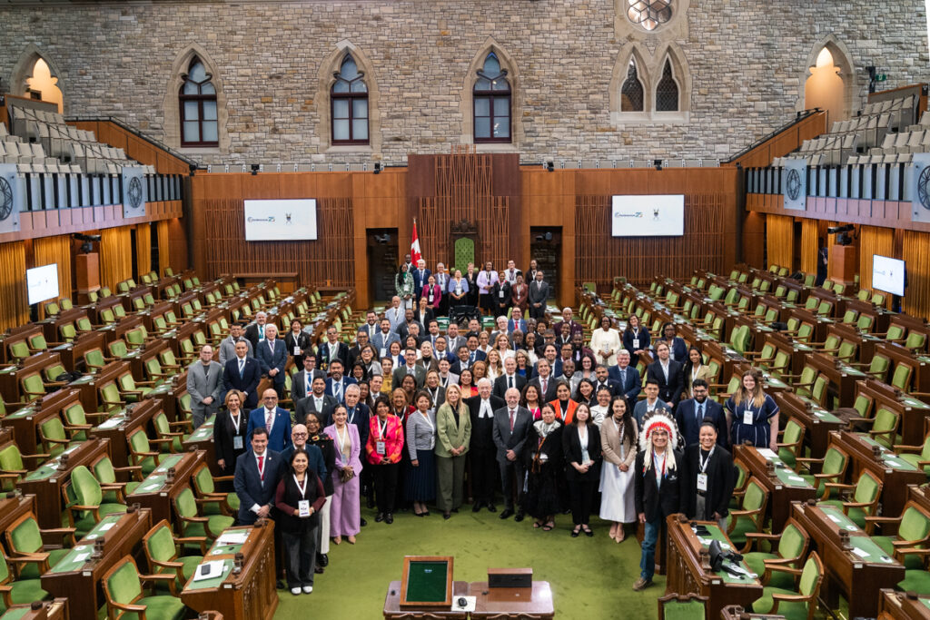 Group photo of a diverse assembly gathered in a grand stone Parliament chamber with green seats and wooden desks.