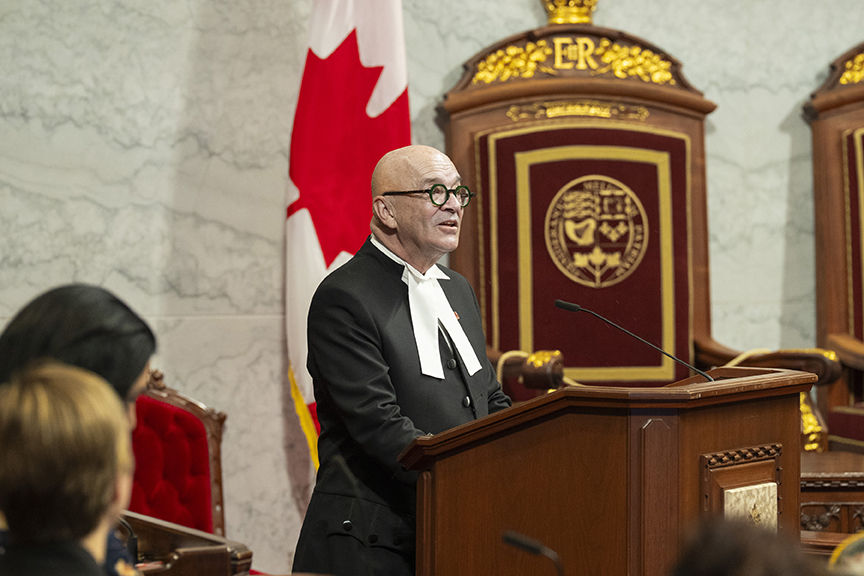 Bald man in a black judicial robe with a white neckband giving a speech at a wooden podium in a formal chamber, Canadian flag visible behind him.