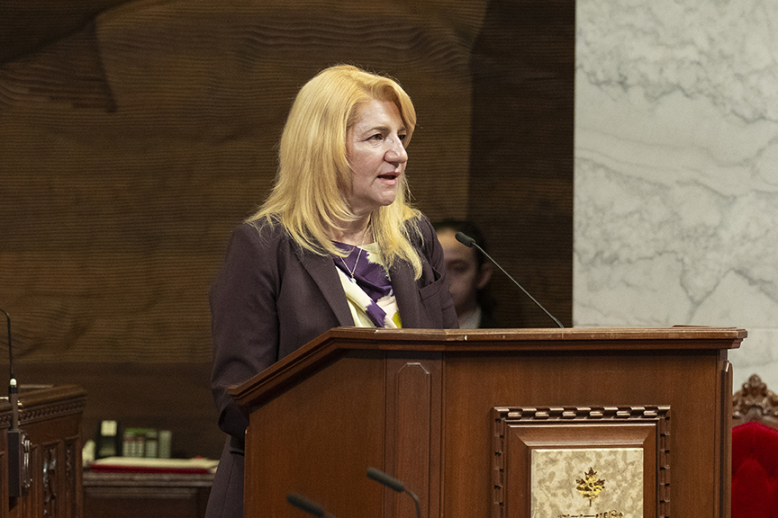 Blonde woman speaks at a wooden podium with a microphone in a formal room with marble wall panels behind her.