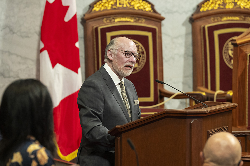 A man in a gray suit speaks at a wooden podium in a formal chamber, with a Canadian flag behind him.
