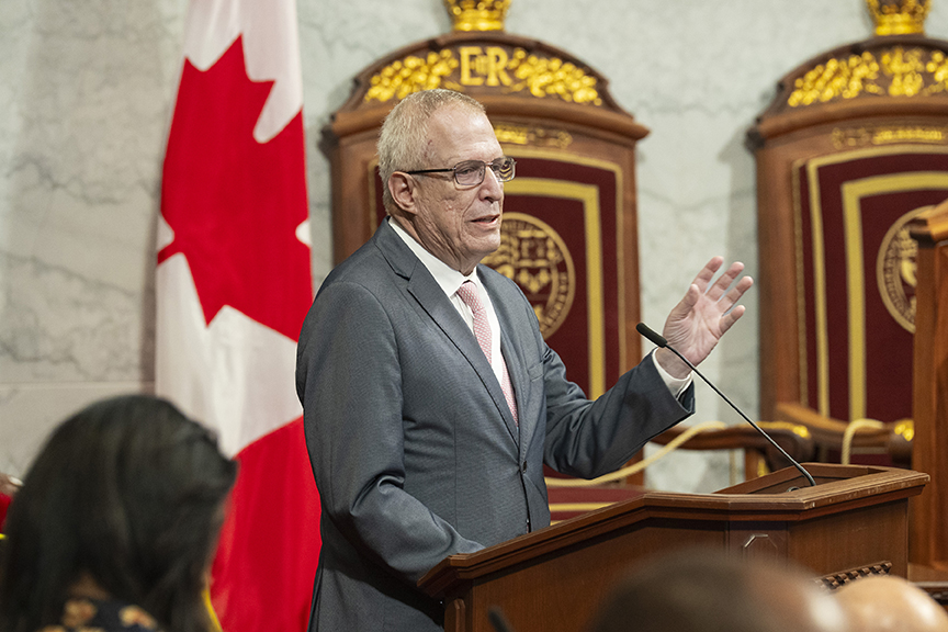 Man in a gray suit and pink tie speaks from a wooden podium in a formal chamber, Canadian flag in background.