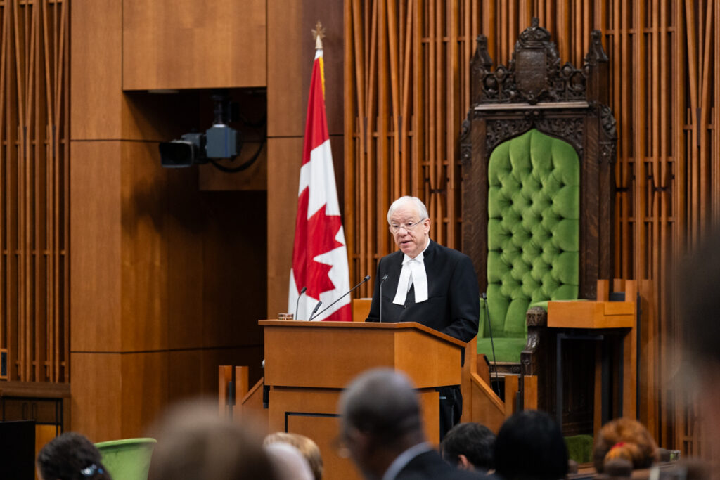 Judge in a black robe speaks at a wooden podium in a courtroom, with a Canadian flag nearby.