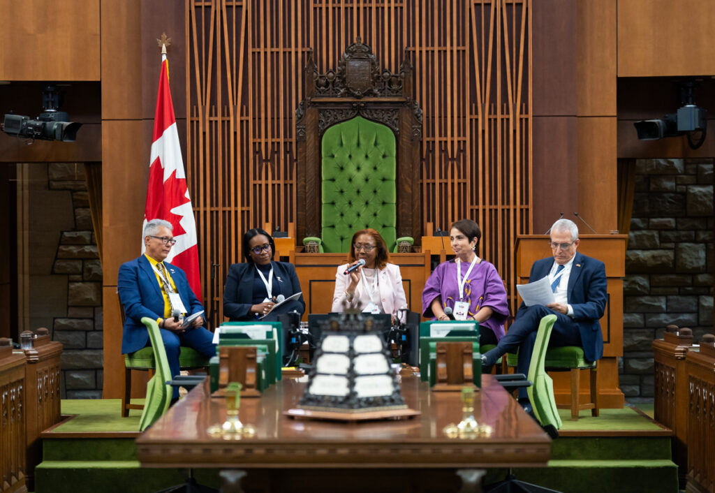 Five panelists seated in a wood-paneled chamber; central speaker holds a microphone, Canadian flag at left.
