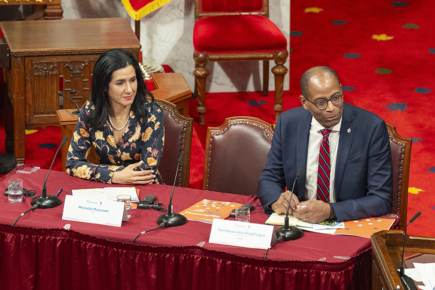 Two officials sit at a red-carpeted panel desk with microphones, nameplates, and documents.