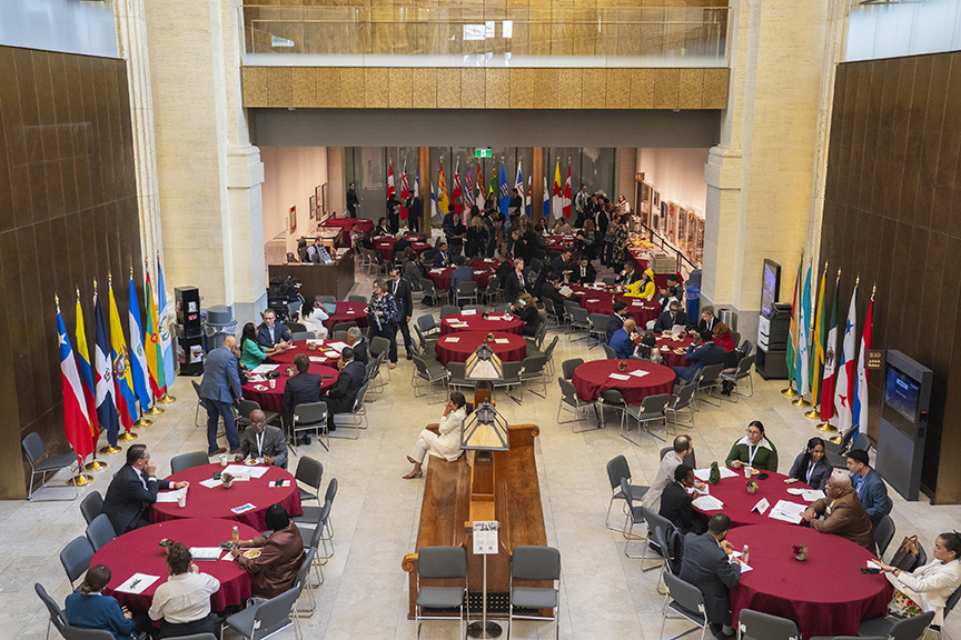 Formal international conference in a large atrium, with many national flags, round tables in red cloth, and attendees surrounding a central podium.