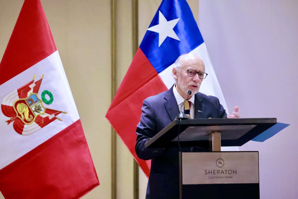 Man in a suit giving a speech at a podium with microphones, flanked by Peruvian and Chilean flags at a Sheraton Lima Historic Center event