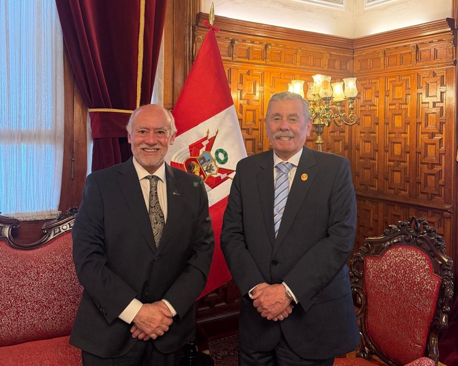 Two men in dark suits smile for a formal photo in a wood-paneled room, with a red-and-white flag behind them.
