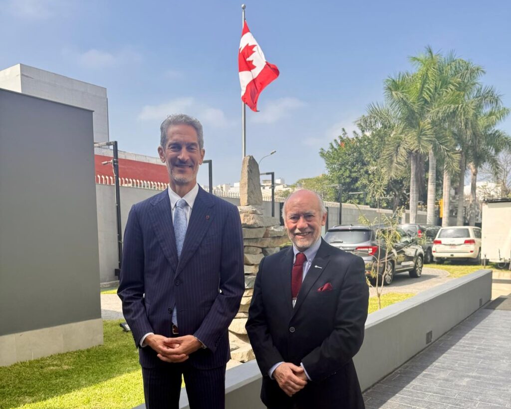 Two men in business suits stand outdoors beside a Canadian flag on a flagpole.