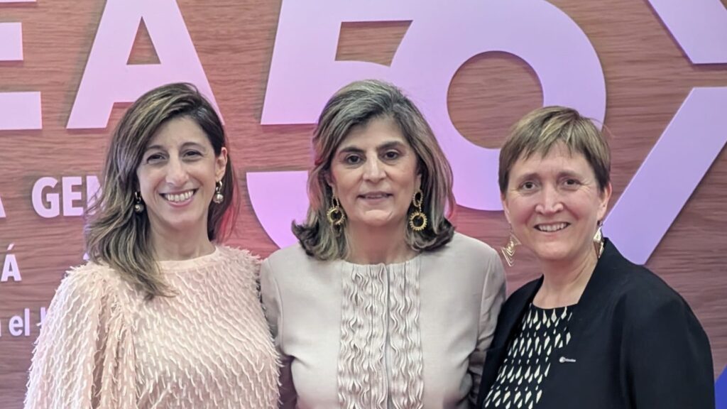 Three women posing and smiling in front of a wooden backdrop with large purple letters behind them, dressed in pastel outfits.