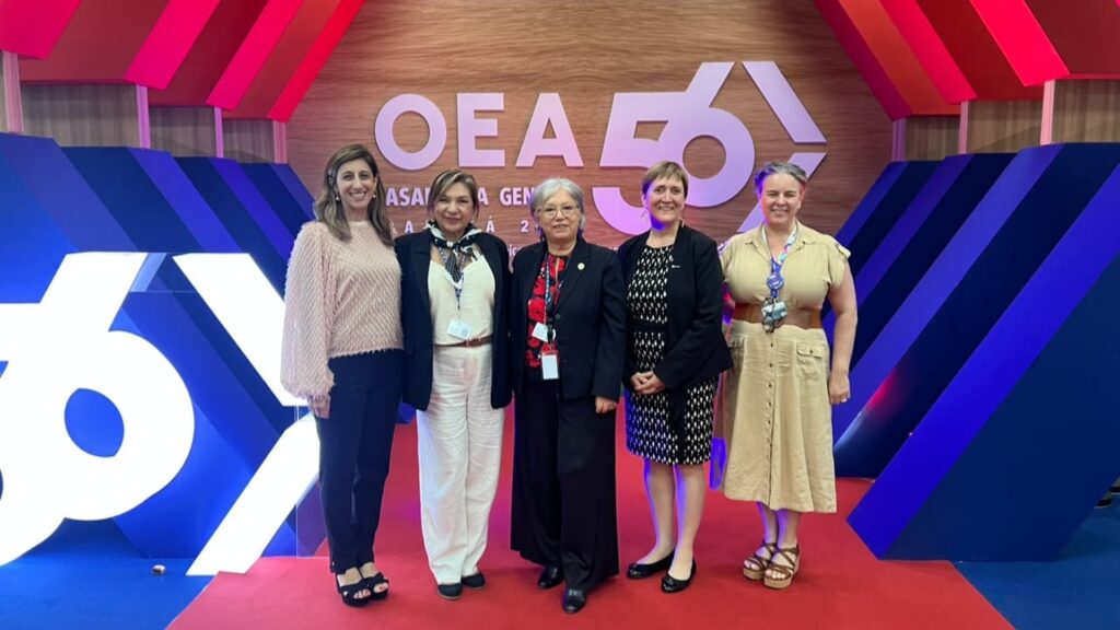 Five women in business attire stand together on a red carpet, posing in front of a colorful event backdrop with a large OEA 65 logo.