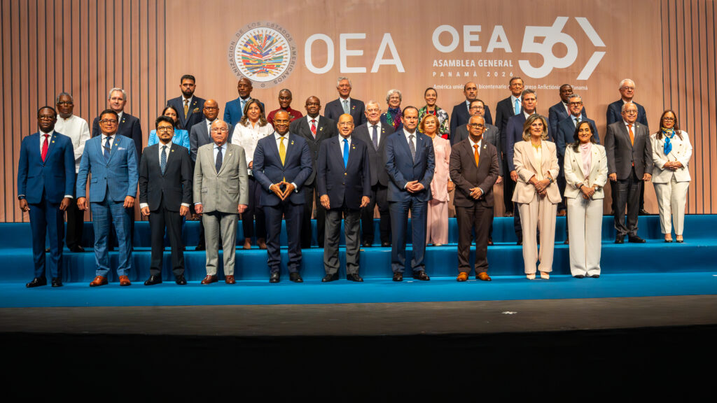 Group of formally dressed officials posing on a blue-carpeted stage during the OEA General Assembly in Panama 2026, with a large banner in the background.
