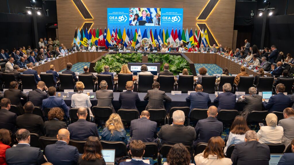 International conference hall with a long U-shaped table of delegates and many national flags in the background, audience watching.
