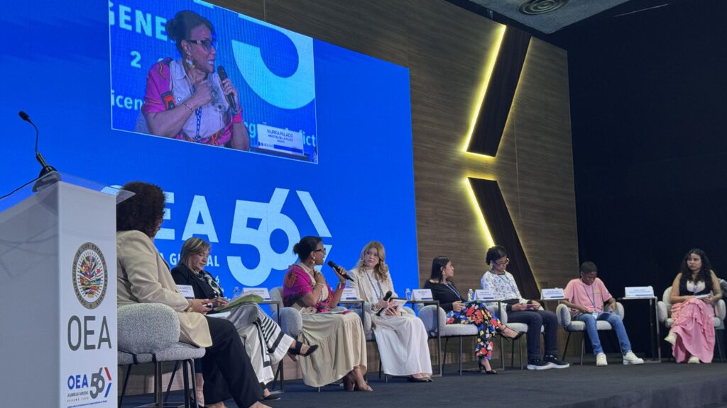 Panel of diverse women seated on stage at a conference, speaking into microphones with a large blue screen behind them.