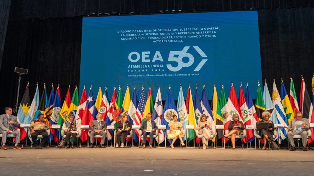 Panel of delegates seated on stage behind a row of national flags at an international summit on a dark stage, blue backdrop.
