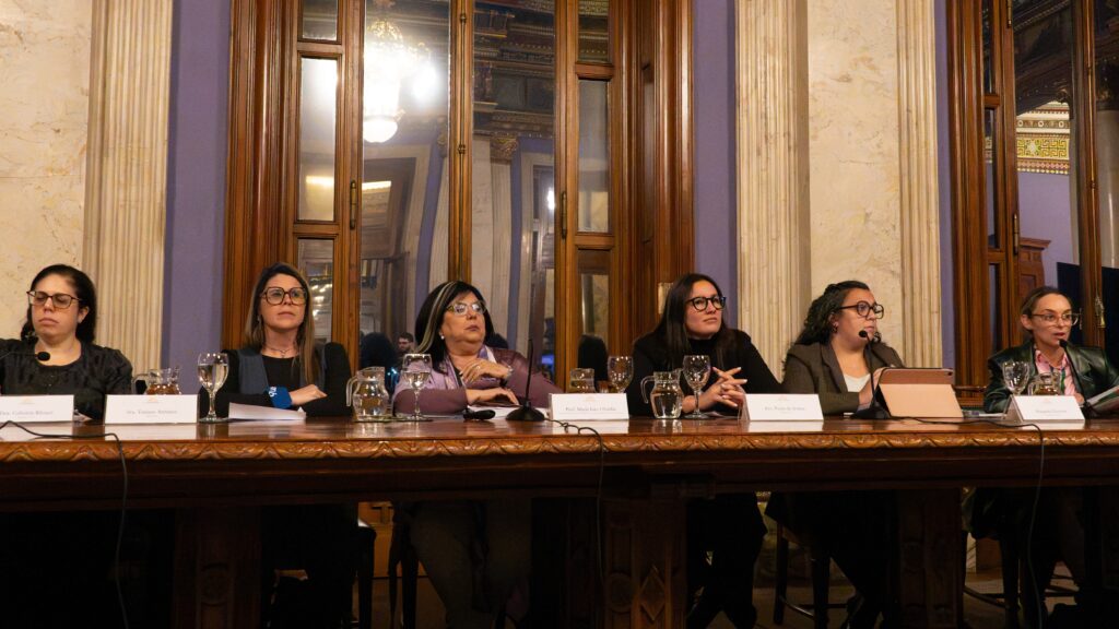 Panel of six women seated at a long ornate table during a formal discussion, with microphones, glasses, and name placards in front.