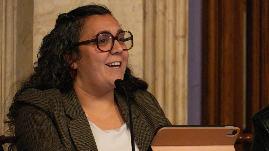 Woman with curly hair and oversized brown glasses speaks into a microphone at a panel, a tablet in front of her.