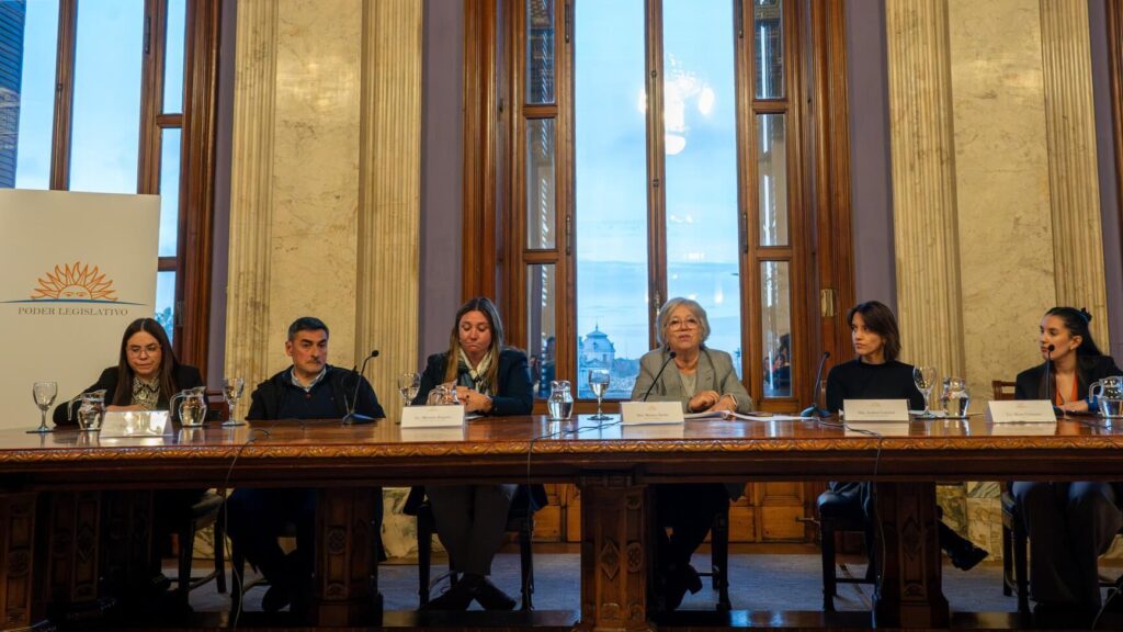 Six panelists sit at a long wooden table in a marble room with tall windows and a city view outside, microphones and nameplates in front of them.