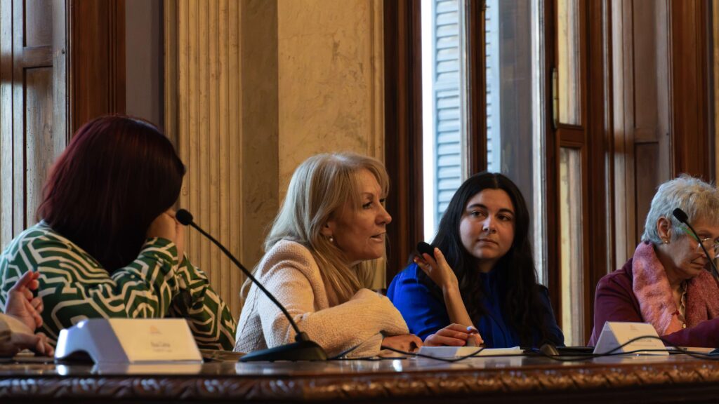 Panel of four women seated at a long table with microphones in a wood-paneled room, mid-conversation during a panel discussion.