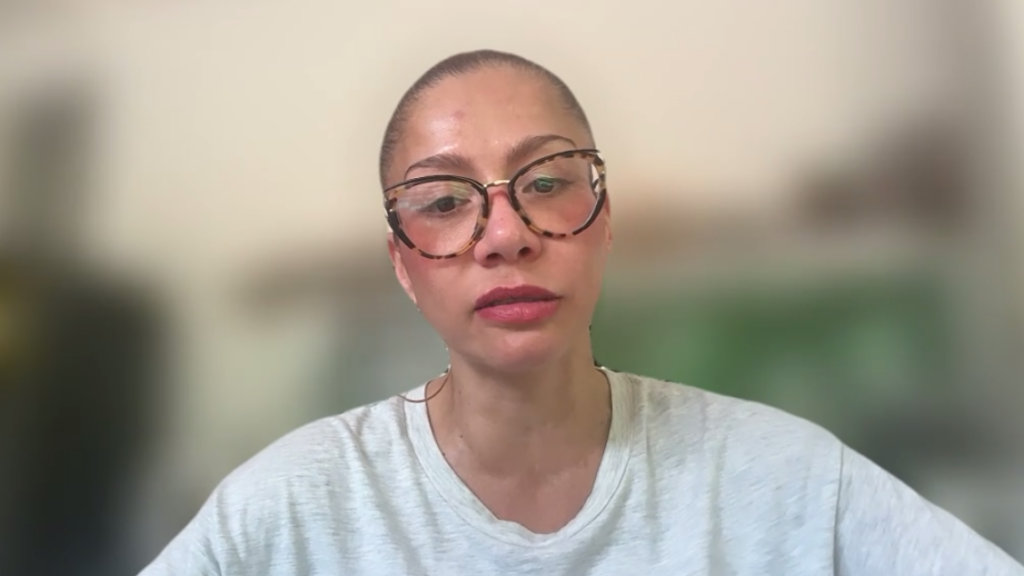 Woman with round tortoiseshell glasses speaking directly to the camera against a blurred indoor background.