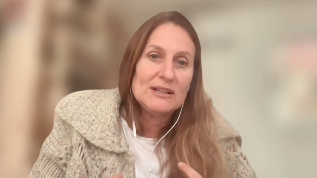 Woman with long brown hair wearing a beige textured coat speaks, wearing white earbuds during a video call or interview.