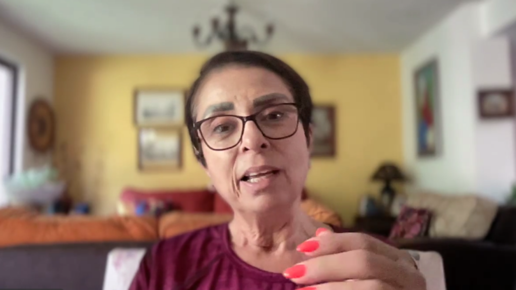 Woman with short dark hair and glasses speaks directly to the camera in a home setting, hand with coral nails visible in foreground.