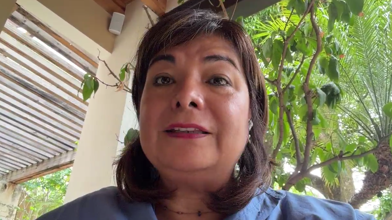Woman outdoors on a porch speaking, with green trees in the background and a blue shirt on.