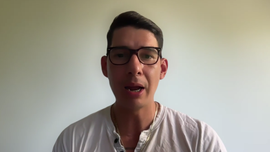 Man wearing glasses and a white shirt speaking directly to the camera against a plain light wall.