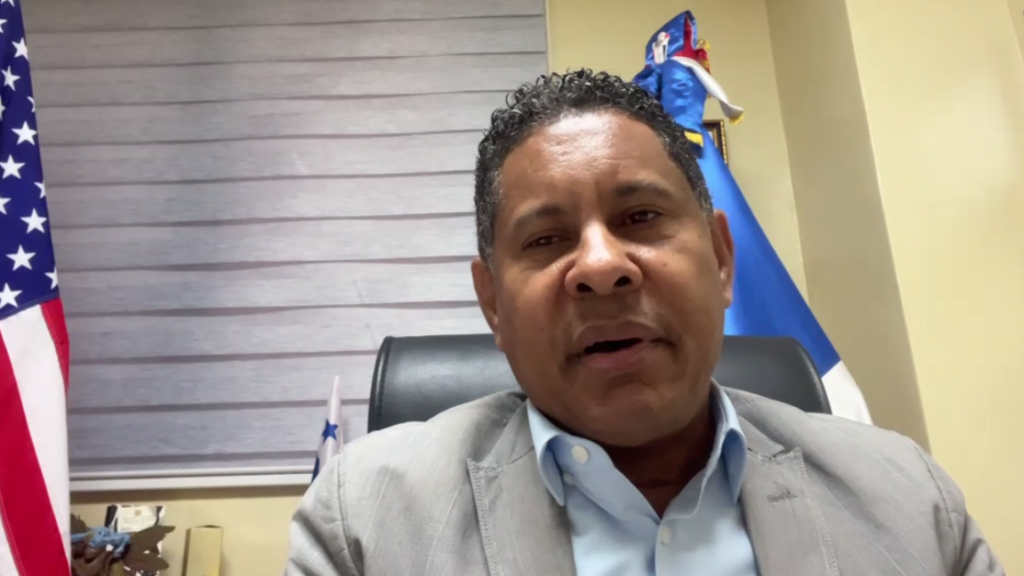 Formal video call or press briefing: a man in a light gray suit and blue shirt speaks from an office with American and other flags in the background.