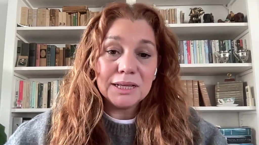 Woman with wavy red hair speaks to camera in a video call, bookshelves and trophies in the background.