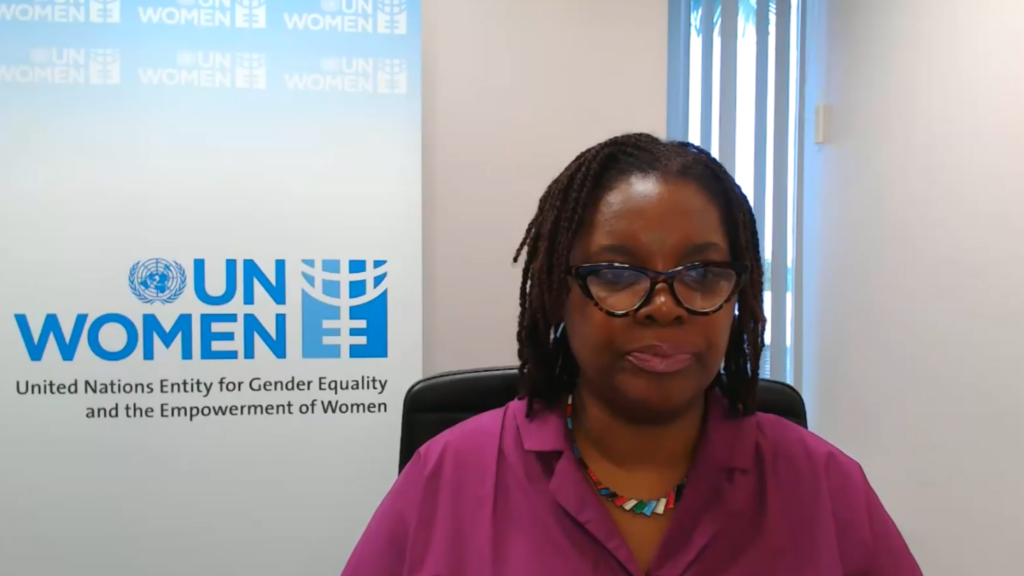 Woman in magenta blouse and glasses seated in an office with a UN Women banner in the background.