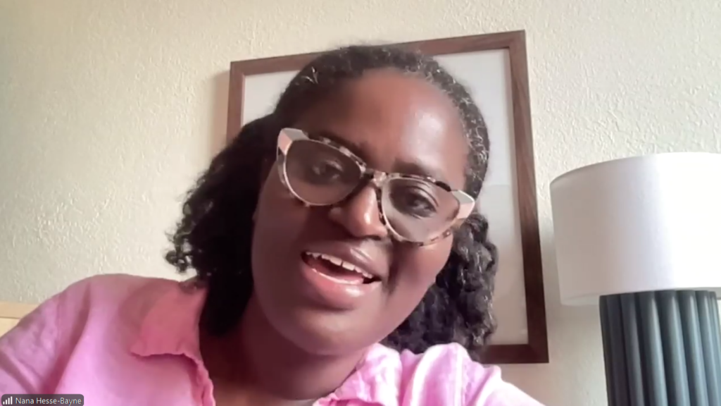 Woman with short curly hair and patterned glasses smiling at the camera during a video call, wearing a pink shirt with a lamp and framed wall art in the background