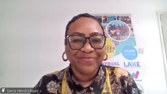 Smiling woman with short hair and glasses on a video call, wearing a patterned shirt, with a colorful banner in the background.