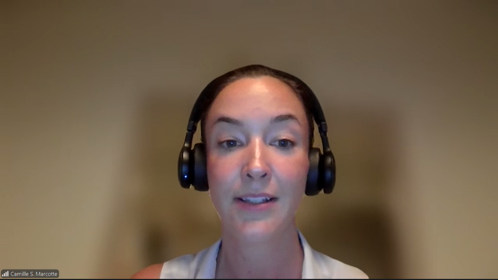Woman wearing a black over-ear headset speaking during a video conference, shown from the shoulders up against a neutral beige background.