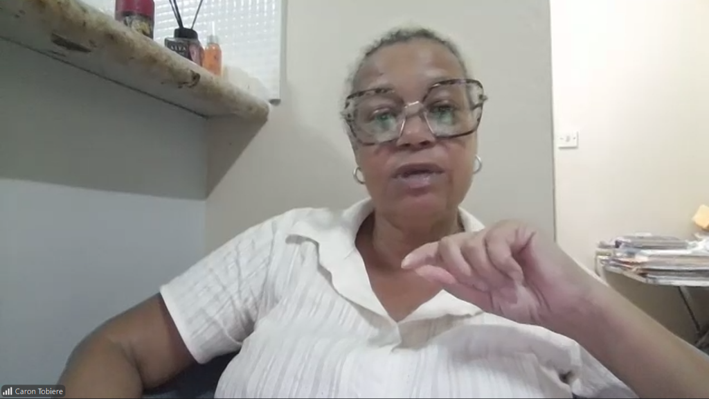 Woman wearing glasses and a white blouse speaks during a video call, gesturing with her right hand in a home setting with a counter and papers in the background.