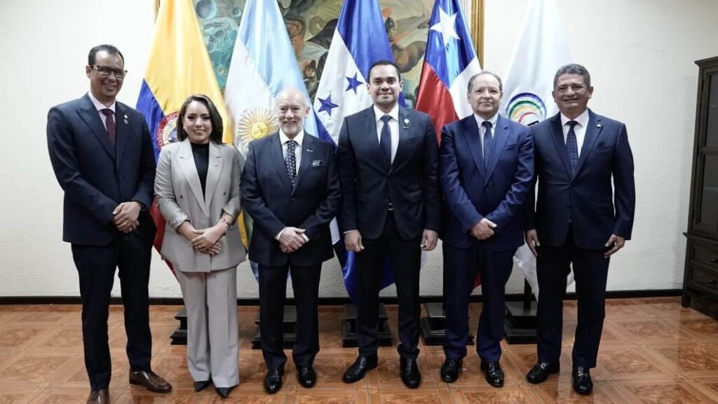 Six formally dressed individuals standing in a line in front of multiple national flags in a formal setting.