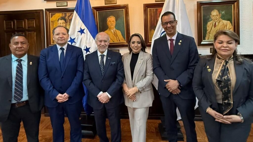 Six professionals pose for a formal group photo in a room with flags and portraits behind them, all in business attire.