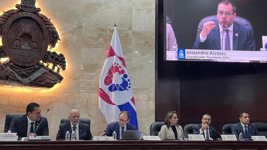 Six officials seated at a long table in a council chamber, with a glowing crest on the wall and a colorful flag behind them.