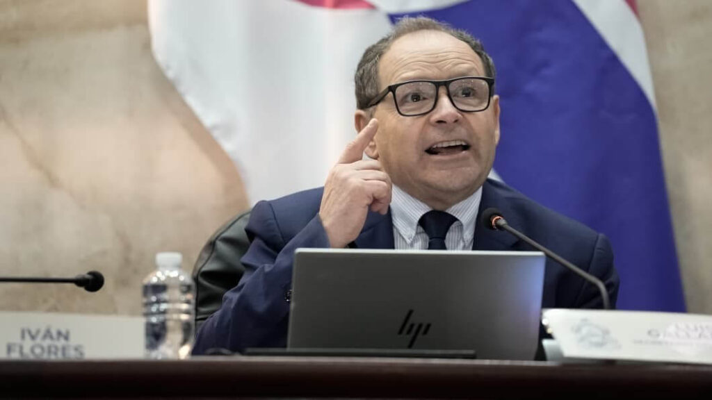 Man in a dark suit and glasses gestures while speaking at a panel, with a laptop and microphone in front of him, Iván Flores nameplate visible.