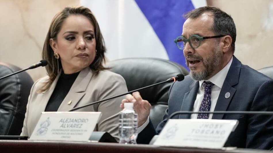 Two officials on a conference panel; a man in a suit speaks into a microphone while a woman in a beige blazer listens beside him.