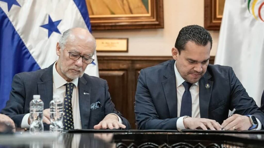 Two men in suits sign a document at a formal meeting, with flags and wood-paneled walls behind them.