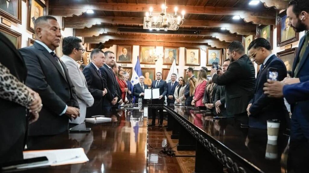 Formal signing ceremony in a wood-paneled room; officials in suits stand along a long conference table as two people present documents at center, with flags and portraits in the background.